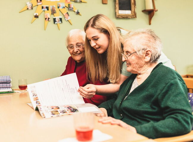 Eine Mitarbeiterin blättert mit zwei Bewohnerinnen durch eine Zeitschrift