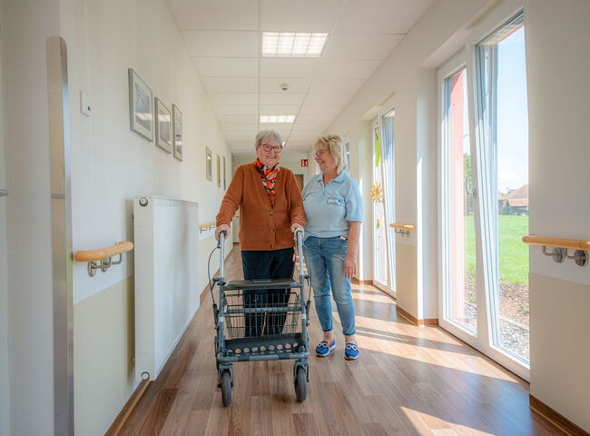 Eine Mitarbeiterin begleitet eine Bewohnerin mit Rollator auf dem Flur