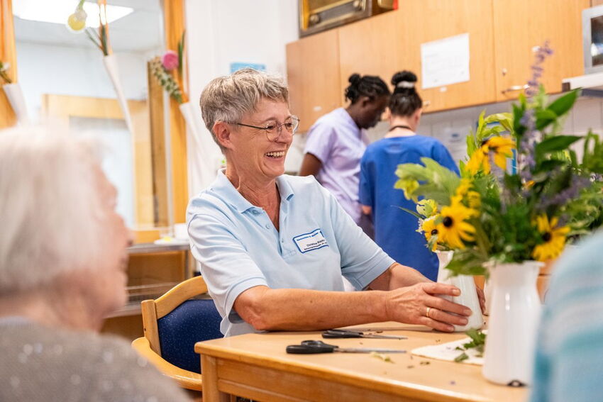 Pflegepersonal mit Blumenstrauß sitzt mit einer Bewohnerin zusammen am Tisch